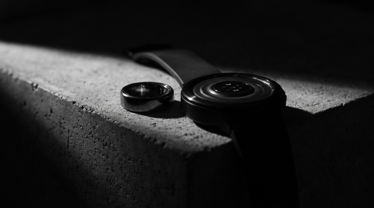 High-contrast black-and-white editorial photo of a smart ring and fitness watch resting face-down on dark concrete