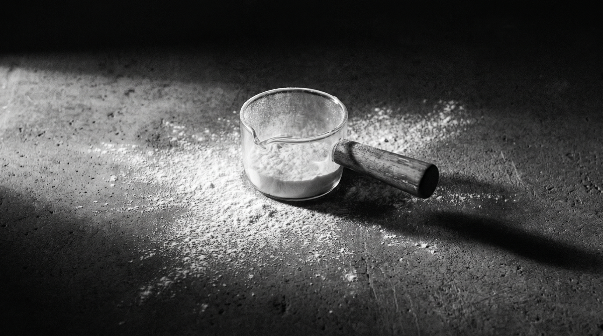 High-contrast black-and-white editorial photo of a glass measuring scoop and scattered protein powder on dark concrete