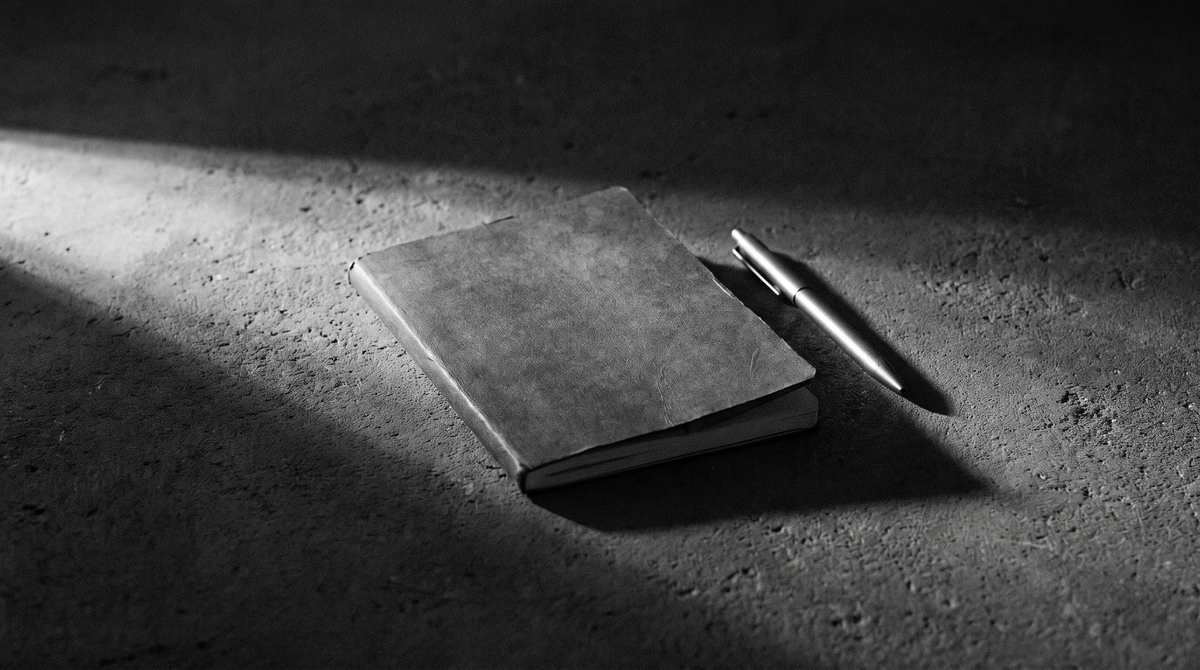 High-contrast black-and-white editorial photo of a closed notebook and pen on dark concrete under harsh side lighting