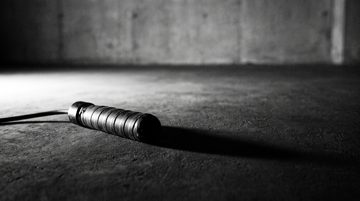 Black and white editorial photograph of a weighted jump rope handle resting alone on dark concrete with dramatic side lighting