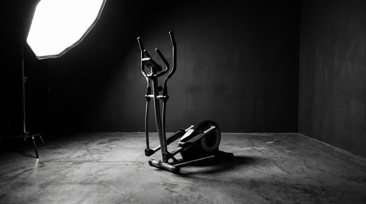 Black and white editorial photograph of an empty elliptical exercise machine in a dark minimal gym studio