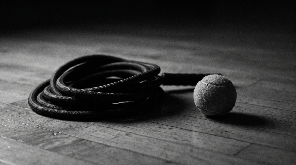 Black and white editorial photograph of a coiled weighted jump rope resting on a wooden gymnasium floor beside a tennis ball, low side angle, cinematic lighting
